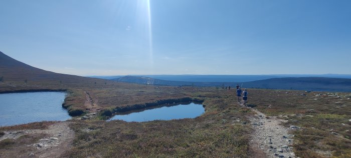 Vandrare på en bergsstig intill en liten sjö med vidsträckta fjällvyer och klarblå himmel.