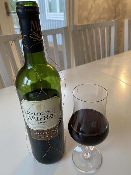 Flaska av Marques de Arienzo Rioja Gran Reserva 2008 bredvid ett glas rödvin på bord.