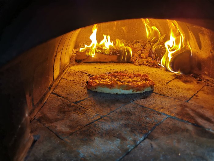 Pizza bakas i en vedeldad ugn med synliga lågor i bakgrunden.