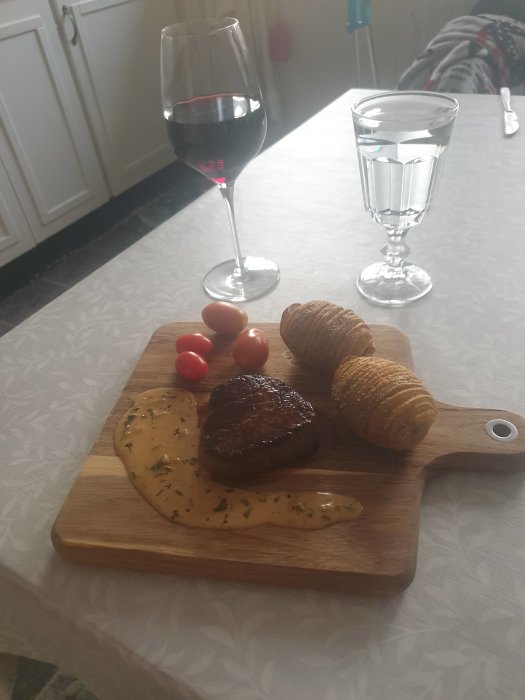 En måltid med biff, hasselbackspotatis, tomater och sås på skärbräda, bredvid ett glas vin och vatten.