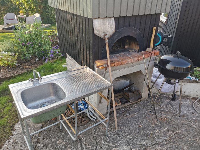 Ett utekök med stenugn, portabelt handfat och kolgrill på en gräsmatta med blommor i bakgrunden.