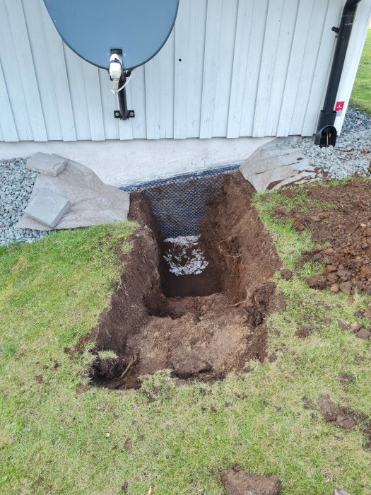 Grävd grop vid husvägg nära kulvert för bergvärme, med synlig dagsljuskabel och satellitantenn ovanför.