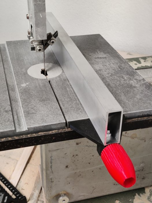 Delvis 3D-utskriven anhåll till en bandsåg med en röd plastdel och aluminiumprofil monterad på sågen.
