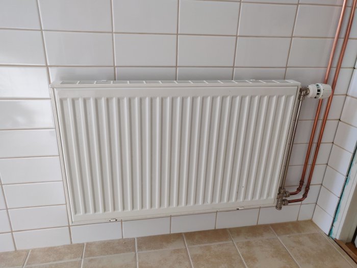 Nyinstallerad vit radiator på kakelvägg med synliga kopparledningar och termostat.