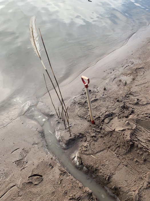 Hemmagjort kanalbygge i sanden vid en strand med ett rödvitt spade och insatta pinnar som markerar kanalen.