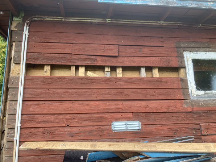 Vägg med rödmålad panel, reglar syns där panel tagits bort, skarvbräda vid ytterkant.