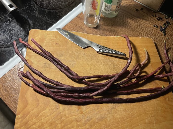 Långa röda bönor av sorten Red Noodle på ett skärbräde med en kniv i bakgrunden.