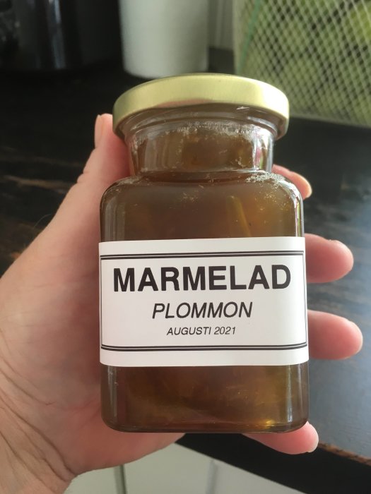 Hand håller ett glasburk med plommonmarmelad och etikett markerad "AUGUSTI 2021".