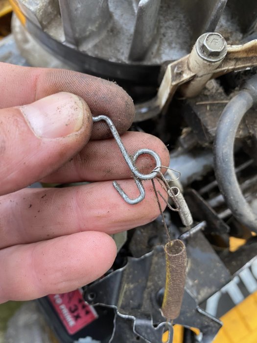 Hand håller en lös, uttänjd fjäder framför en bränsleinställningsmekanism på en motor.