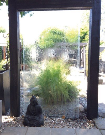 Vattenvägg i trädgård med Buddha-statyn och svarta dekorstenar, synligt genom pergolans öppning.