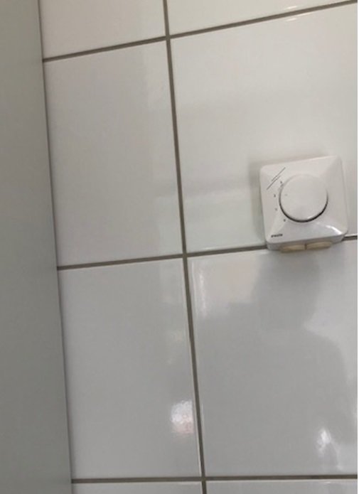 Vägg med vita kakelplattor och en varvtalsregulator till ventilationsanläggning.