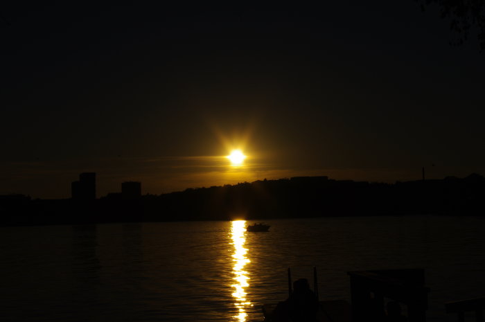 Solnedgång över en vattendrag med en liten båt och stadsilhuett i bakgrunden.