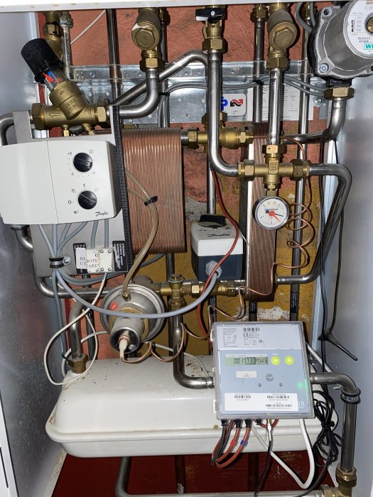 Golvvärmepump och fjärrvärmecentral i teknikrum, rörsystem och mätare synliga.