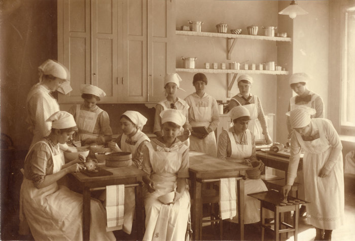 Historiskt kök med kvinnor som förbereder mat vid fristående bord och får instruktioner.