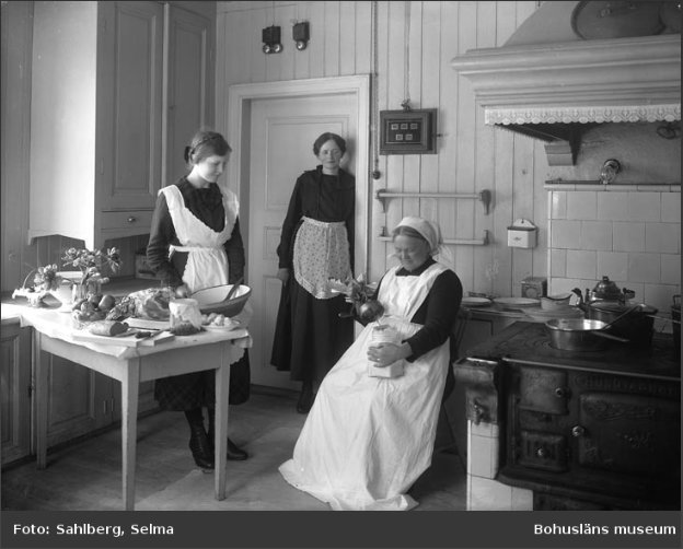 Kvinnor i historiskt kök med fristående bord, stora kopparkärl och låg vedspis.