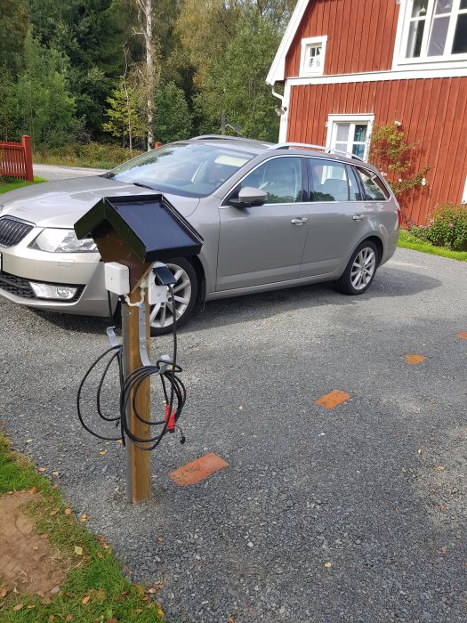 Hemmagjord svart laddningsstation för elbil framför en grå bil och rött hus, troligt DIY-projekt.