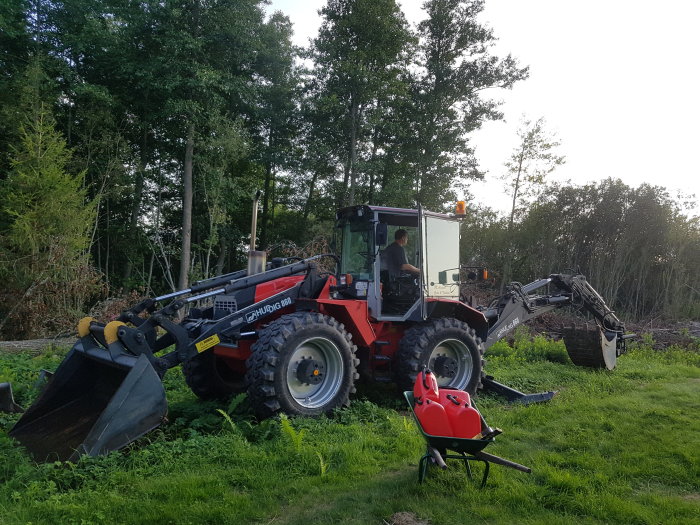 Röd traktor med frontlastare och grävmaskinstillbehör i ett grönt fält med skog i bakgrunden.