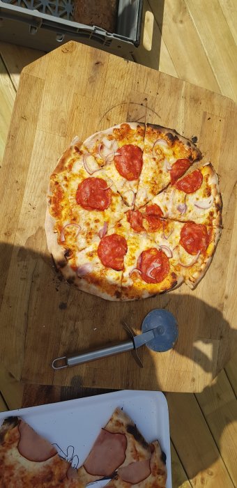 Hembakad pizza med pepperoni och lök på en träskärbräda utomhus, pizzaugn i bakgrunden.