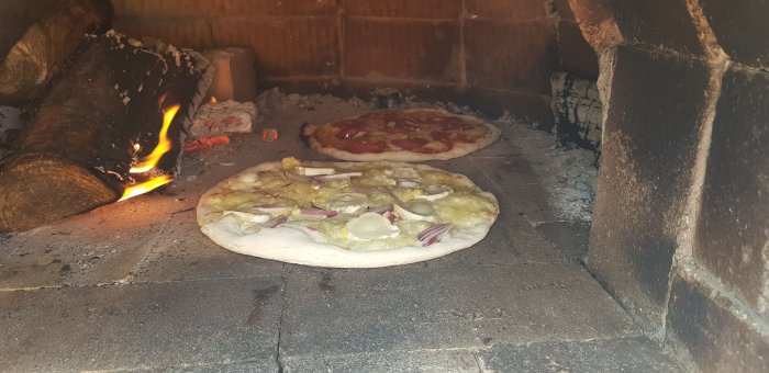 Två pizzor gräddas i en vedeldad pizzaugn med synliga lågor och vedträn.