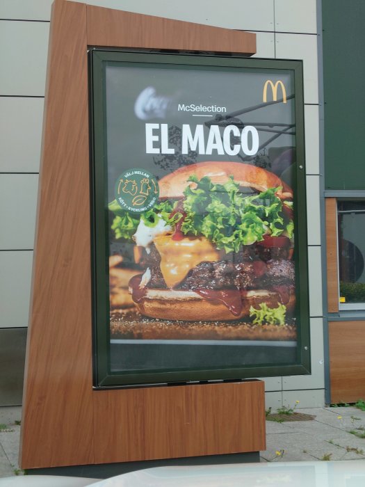 Reklamskylt för McDonald's El Maco burgare med sovs och sallad på trästolpe.