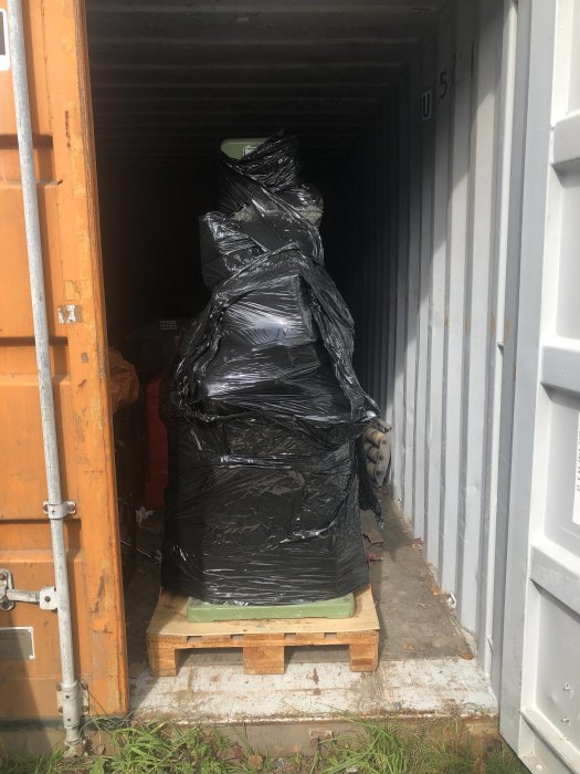 Föremål inslaget i svart plast på pall väntar på transport i en öppen fraktcontainer.