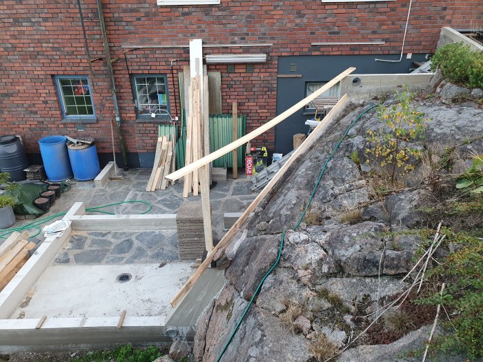 Byggplats med bräda lutad mot husvägg, verktyg och byggmaterial förberedda för gjutning av betongfot.