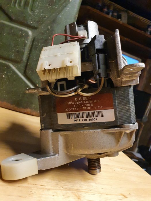 Eldmotor från en demonterad tvättmaskin med etikett och kabelanslutningar på en verkstadsbänk.