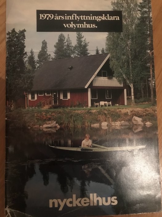 Rödfärgat Nyckelhus vid sjökant med skog i bakgrunden och person som fiskar i en båt på vattnet.