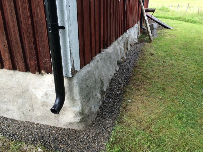 Renoverad husgrund med ny betong och dränering med grus vid röd trävägg och stuprör.