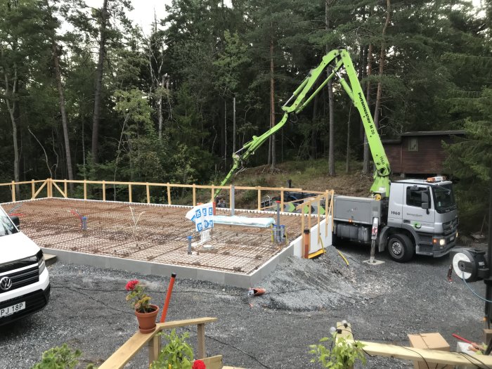 En byggarbetsplats med gjutet bjälklag och armeringsjärn, mobilkran som arbetar och byggmaterial runtomkring i en skogsmiljö.