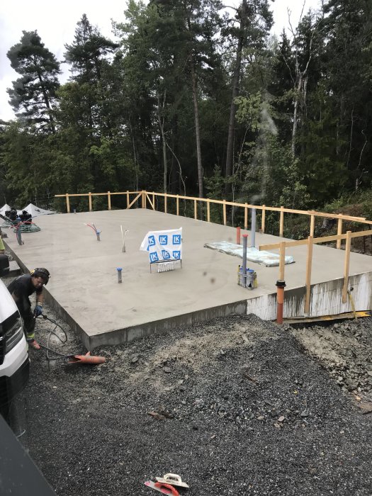 Nyligen gjuten betongplatta för ett husbygge omgiven av skog med uppstick och verktyg synliga.