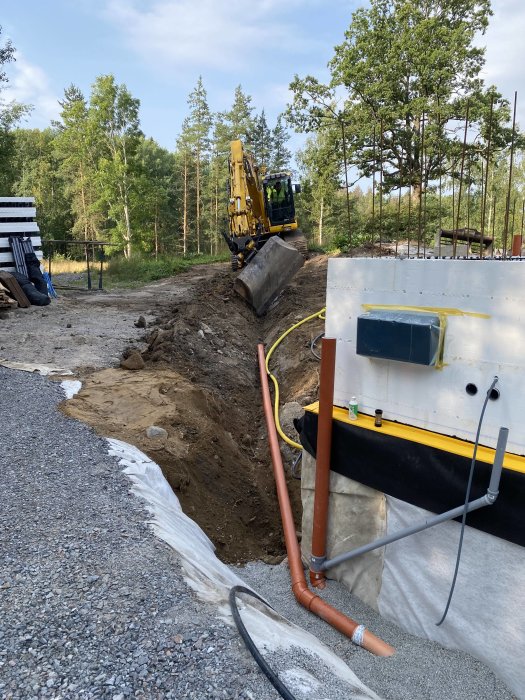 Grävmaskin vid byggarbetsplats, återfyller grunden med schaktmaterial, synliga rör och isolering i förgrunden.