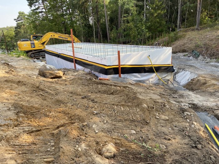 Armerad betongplatta på byggarbetsplats med grävmaskin och återfylld källare i skogsmiljö.