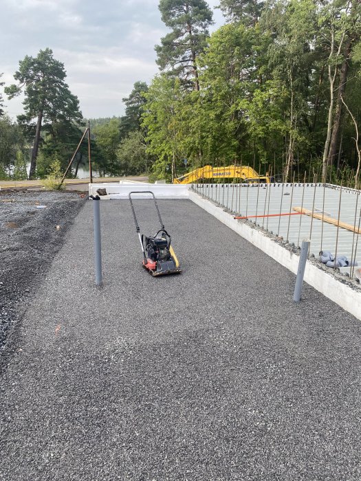 Nybyggd betonggrund med armeringsjärn och nylagd makadam, omgiven av skogsnatur.