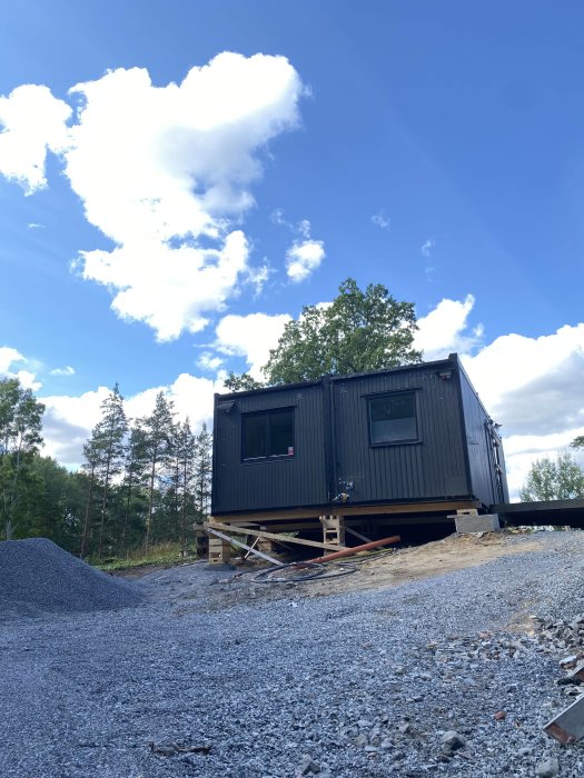 Byggbodar på tomten som används till bostad under ett byggprojekt, omgivna av grus och skog.