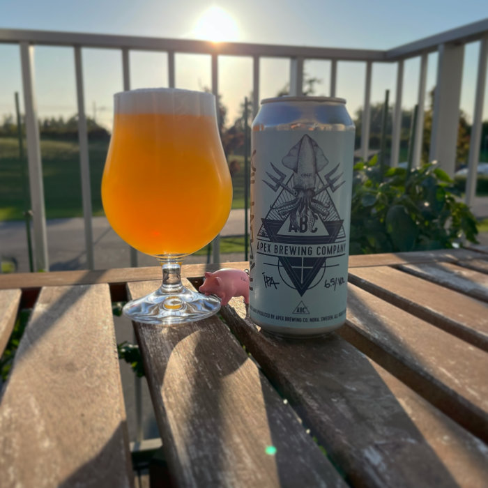 Ölglas fyllt med orangefärgad dryck på träbord bredvid en burk från Apex Brewing i solnedgång.