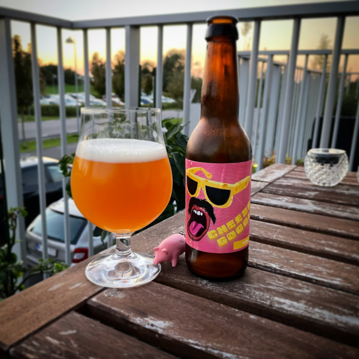 Ölflaska och glas fyllt med öl på träbord, med en balkong och skymning i bakgrunden.