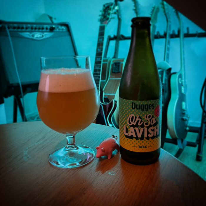 Ett glas öl med skum vid sidan av en Dugges ölflaska och en liten rosa leksaksgris, med gitarrer i bakgrunden.