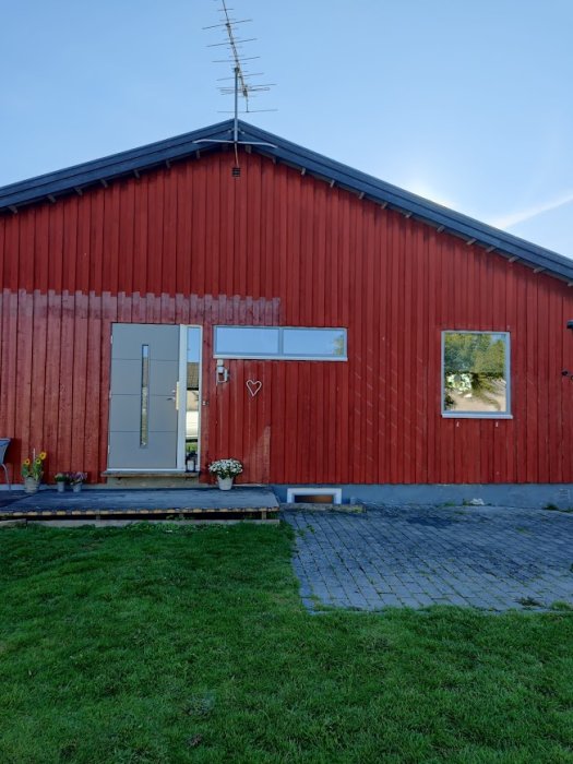 Röd träfasad på hus utan fönsterfoder, grön gräsmatta och grå stentrappa.