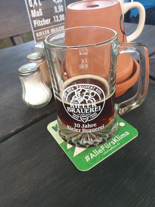 Ölsejdel med logotyp för Kieler Brauerei, halvfull, på ett bord med saltkar och reklammaterial i bakgrunden.