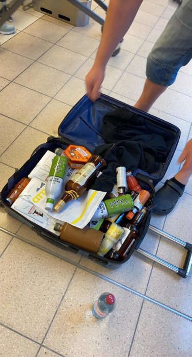 Öppen resväska på golv fylld med olika flaskor av alkoholhaltiga drycker och kläder.