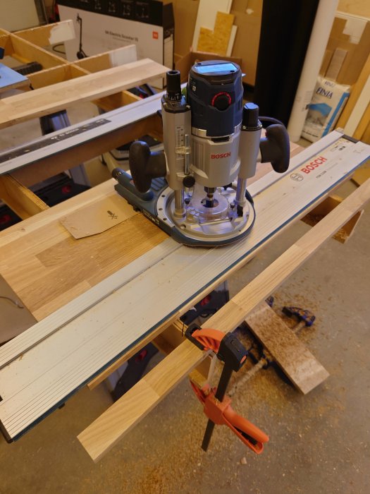 En överfräs från Bosch placerad på en vitpigmenterad träplatta som är fastklämd på ett arbetsbord.