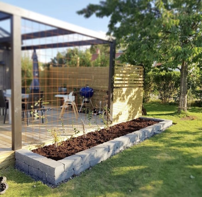 Nyplanterad trädgårdsrabatt med stenkanter framför en pergola med sittgrupp och grill.