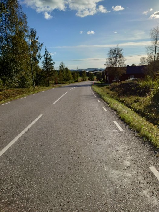 Asfalterad landsväg med vitmarkerade körfält, omgiven av träd och en röd stuga, under klar himmel.