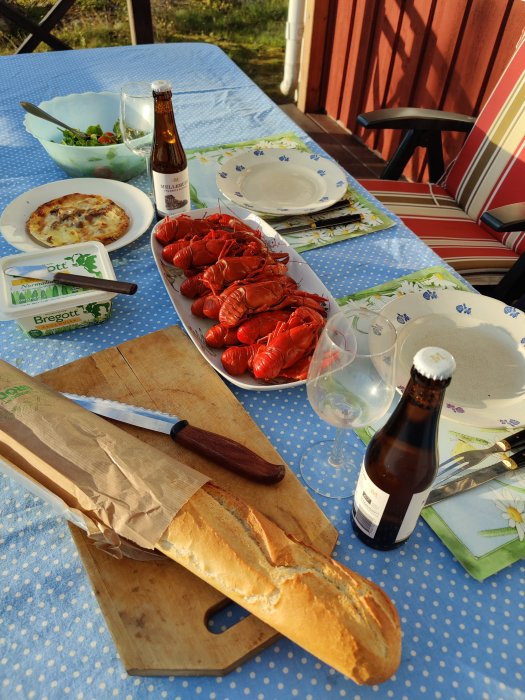 Sommarbuffé utomhus med kräftor, paj, bröd, sallad, smör och öl på ett dukat bord.