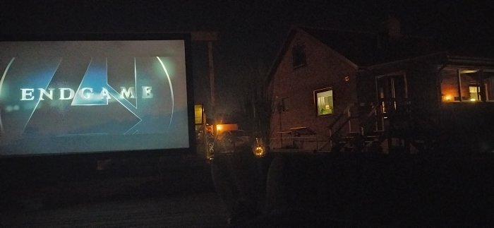 Utomhusbiograf med stjärnklar himmel, projicerad film "ENDGAME" bredvid ett bostadshus på kvällen.