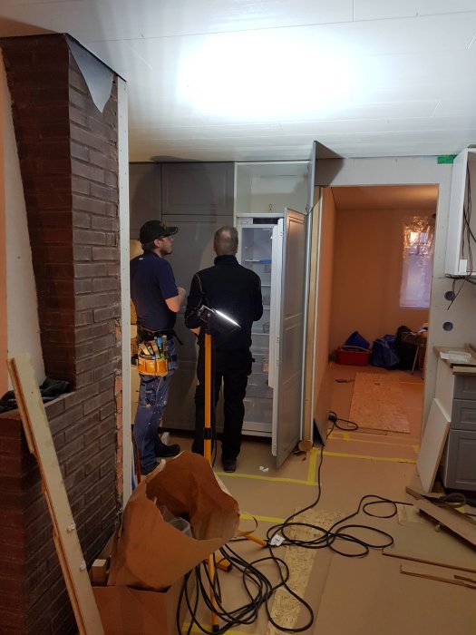 Två män installerar en vitvara i ett kök under renovering, med verktyg och material synliga.