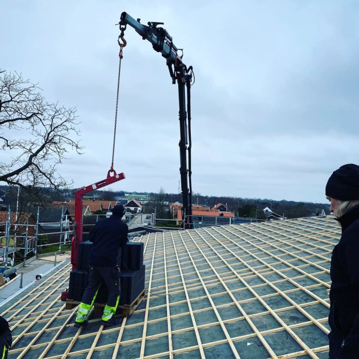 Arbetspersonal använder kranbil för att lyfta en pall med byggmaterial vid takläggning.