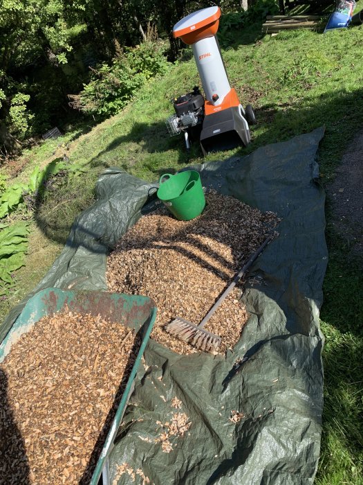 Flismaskin med en hög av flisat grenmaterial, skottkärra och kratta på en presenning.