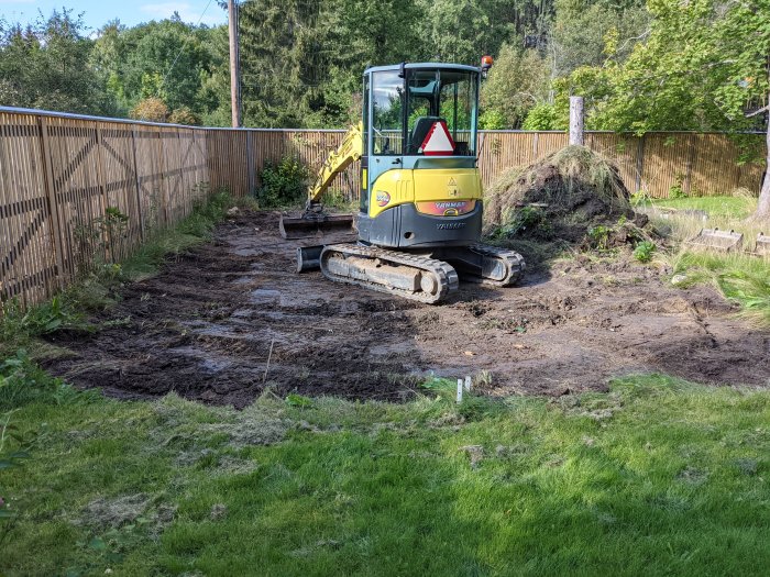 Minigrävare på lerig arbetsplats i trädgård efter grävningsarbete.